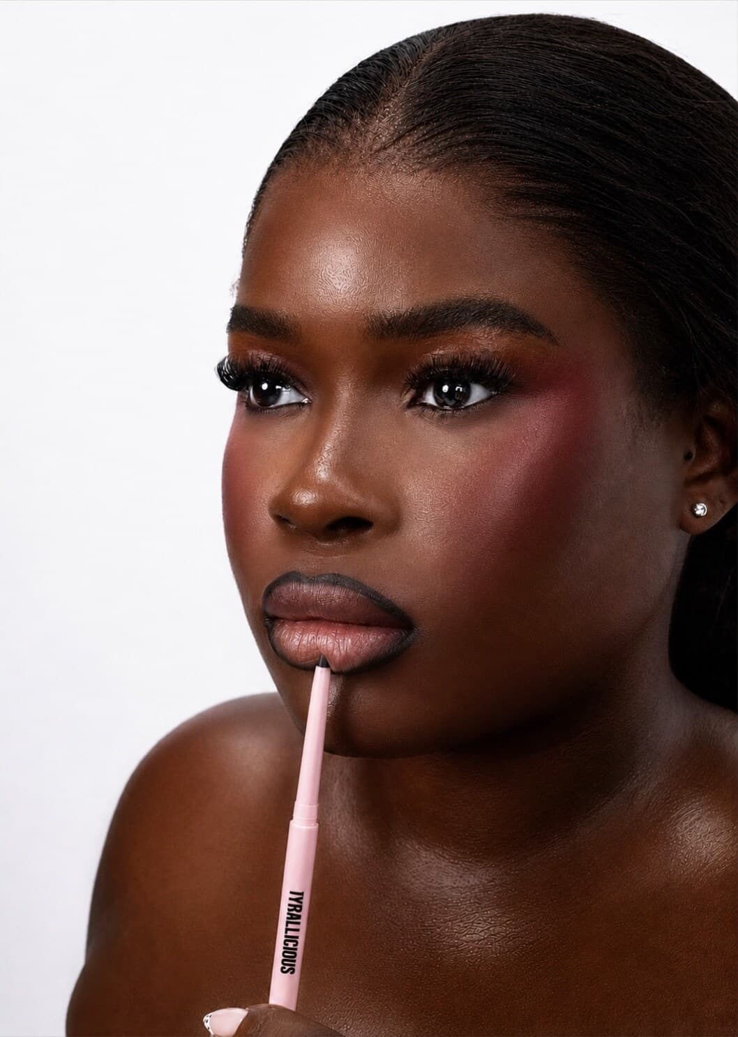 Model with deep skin holding an Iyrallicious lip liner at the lower lip.